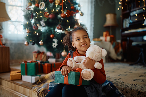 Portrait of smiling African American little girl with teddy bear and gift box on Christmas looking at camera.