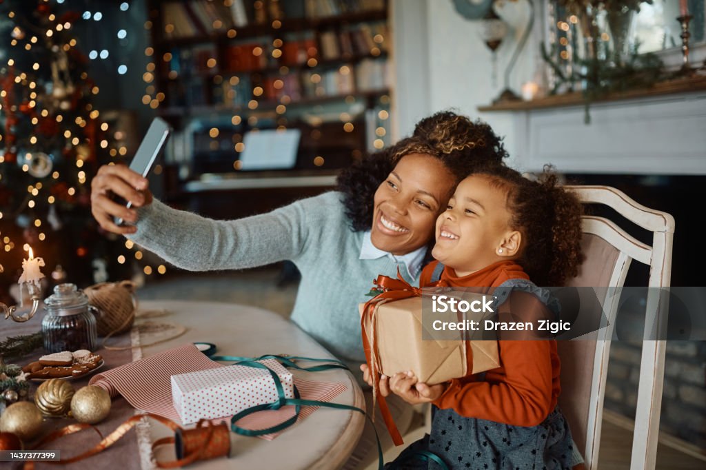 Cheerful black mother and daughter taking selfie on Christmas day at home. Happy African American mother and daughter having fun while wrapping Christmas presents and taking selfie with cell phone at home. Gift Stock Photo