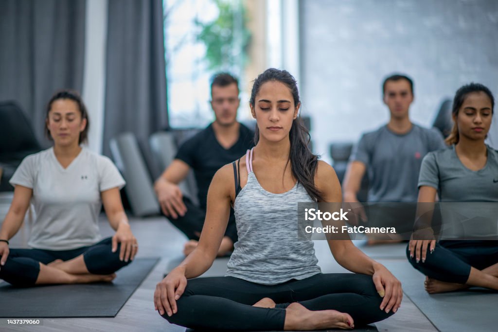 Woman Meditating A young woman of Middle Eastern decent sits in a Meditation class with her hands resting on her knees and her eyes closed.  She is dressed comfortably in athletic wear and focusing on her breathing as she meditates with a group of peers in a class. 18-19 Years Stock Photo