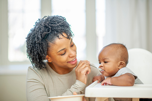A young mother of African decent sits beside her infant son as he sits in his highchair and she feeds him his morning cereal.  She is dressed casually as she holds the bowl in one hand and the spoon in the other.