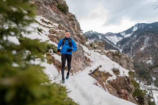 Active passionate trail runner in his 50's taking a run on a snowy mountain path. He is wearing warm clothes for outdoor sport activities and carrying a backpack.
