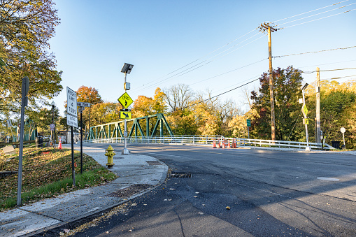 Recent construction activity and equipment is still evident at the site of a modernized road intersection. There is brand new solar-powered pedestrian crosswalk flashing lighting equipment and signage; and work is almost complete on the refurbished bridge infrastructure and highway road surface crossing over the Erie Canal (New York State Barge Canal). Late October autumn morning sunrise in the village of Pittsford, NY - near Rochester, New York.