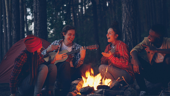 young man tourist is playing the guitar while his friends are singing and laughing sitting around fire in the wood in the evening enjoying nature and good company.