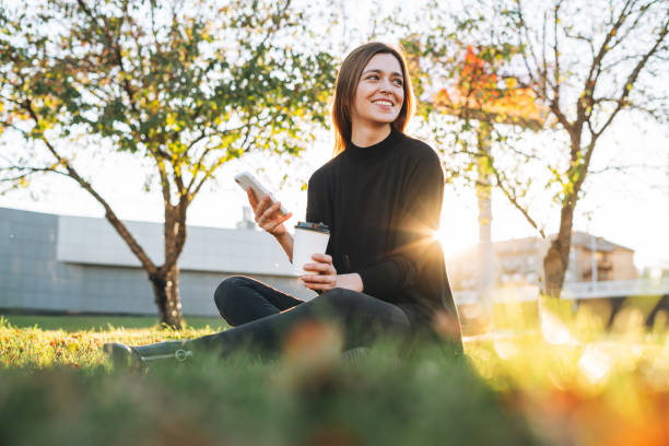 portret młodej studentki z długimi włosami za pomocą telefonu komórkowego w parku miejskim w złotej godzinie - entrepreneur lifestyles nature environment zdjęcia i obrazy z banku zdjęć