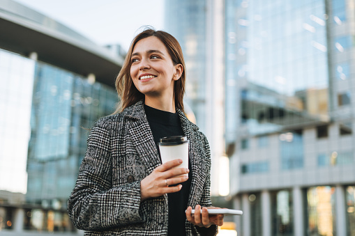 Young smiling woman in coat with coffee cup using mobile phone in evening city street