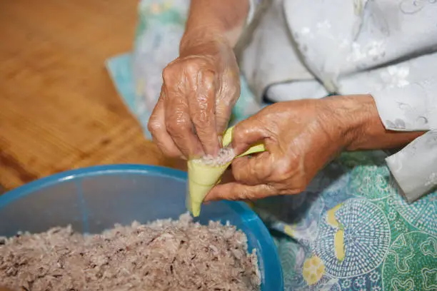 Photo of Hands of senior woman wrapping the sticky rice with palm leaf or Ketupat Palas