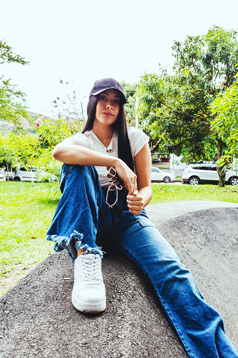 latin girl sitting trendy in jean and cap in outdoor park happy, smiling.