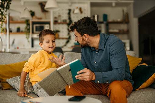 A mid-aged father with a small son sitting at the sofa by the table indoors, making homework. Father holding a book and helping his son with a reading.
