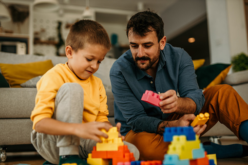 Caring young single caucasian father helps cute kid son play on a warm floor together, happy family dad and little child boy having fun building a constructor tower from colorful blocks.