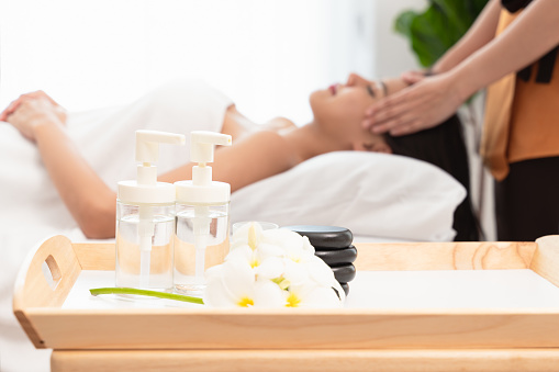 Spa composition with essential oil bottles, stones and frangipani flower. Blurred figures of young woman enjoying facial massage and masseur in background.