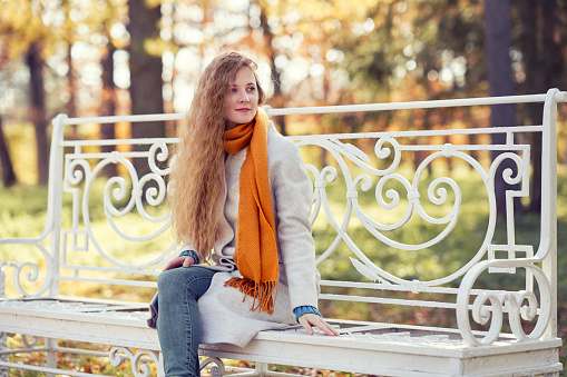 A young red-haired woman is sitting on a bench in an autumn park. A girl in a white coat is resting in the autumn forest