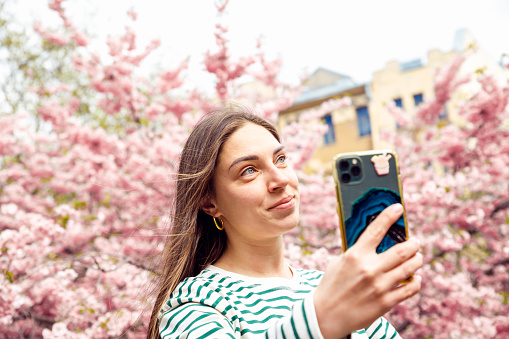 Smiling Young Caucasian woman blogger making selfie with smartphone. Female outdoors against cherry blossom sakura is taking self portrait with mobile phone. Summer Spring headshot lifestyle portrait.