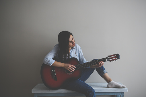 serious caucasian woman with long hair plays a guitar on a table against the wall in a real interior