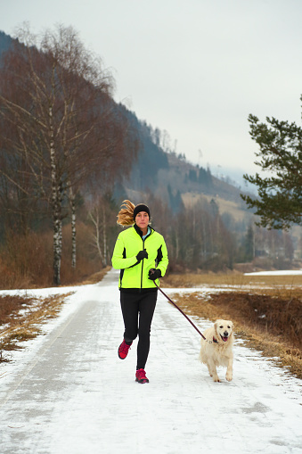 Woman in sports clothing running in winter. Her dog running with her. Snow on road and fog in the park.