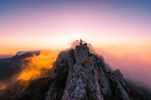 An aerial shot of a female with hands up on the top of the mountain at sunset