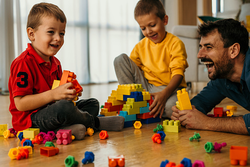 Happy millennial 30s dad playing with his two little sons and helping them to build toy towers from construction plastic blocks. Joyful kid playing learning games with daddy on the heating floor