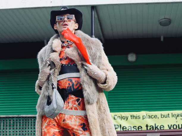 LGBTQ. Person, wearing orange with red fan and cowboy hat. Art photo, modeling photo, sunglasses, leather, wild, beautiful, fashion. Freedom. Chains. Green, jeweled gloves, purse, building in background, outdoors.