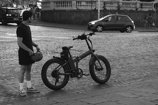 Napol, Italy – July 29, 2022: A grayscale of a young man holding a helmet and standing next to a bicycle in Naples, Italy