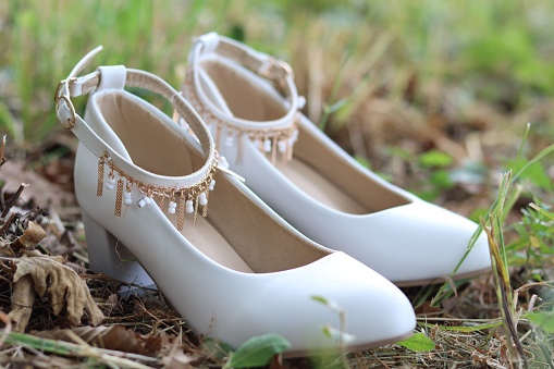 White wedding shoes laid in the grass, with a low heel, jewelry on the ankle strap.