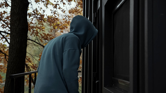 Thief in dark clothes with hood looks around and looks into house through door glass. Man picks door lock and enters house on autumn day side view