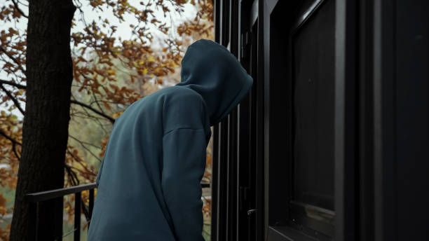 Thief in dark clothes picks door lock and enters house Thief in dark clothes with hood looks around and looks into house through door glass. Man picks door lock and enters house on autumn day side view intercepting stock pictures, royalty-free photos & images