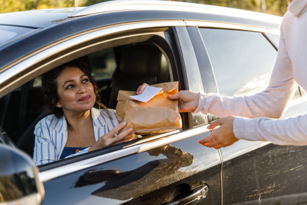 Delivery woman at the curbside pickup giving takeaway order to a customer Cut out shot of smiling mid adult woman sitting in driver's seat in her car and receiving a paper bag with her takeaway order, from the service woman, at the curbside pickup. curbsidepickup stock pictures, royalty-free photos & images