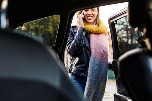 Selective focus shot of smiling young blond woman entering a car through the back door and talking on the phone with a friend.