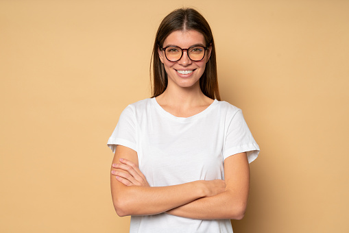 Portrait of successful confident student girl in white mockup t-shirt with copy space for your advertising content and stylish glasses standing with crossed hands on brown studio background