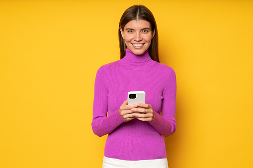 Student girl in pink turtleneck using learning app to study foreign languages holding smartphone on yellow studio background, looking at camera with smile