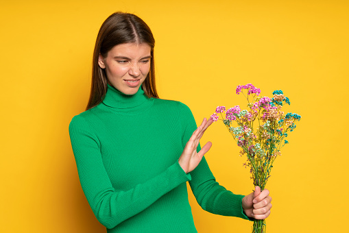 Portrait of displeased woman holding bouquet of flowers doing no gesture, posing on yellow studio background with ugh facial expression because of allergy to pollen or disgusting flower smell