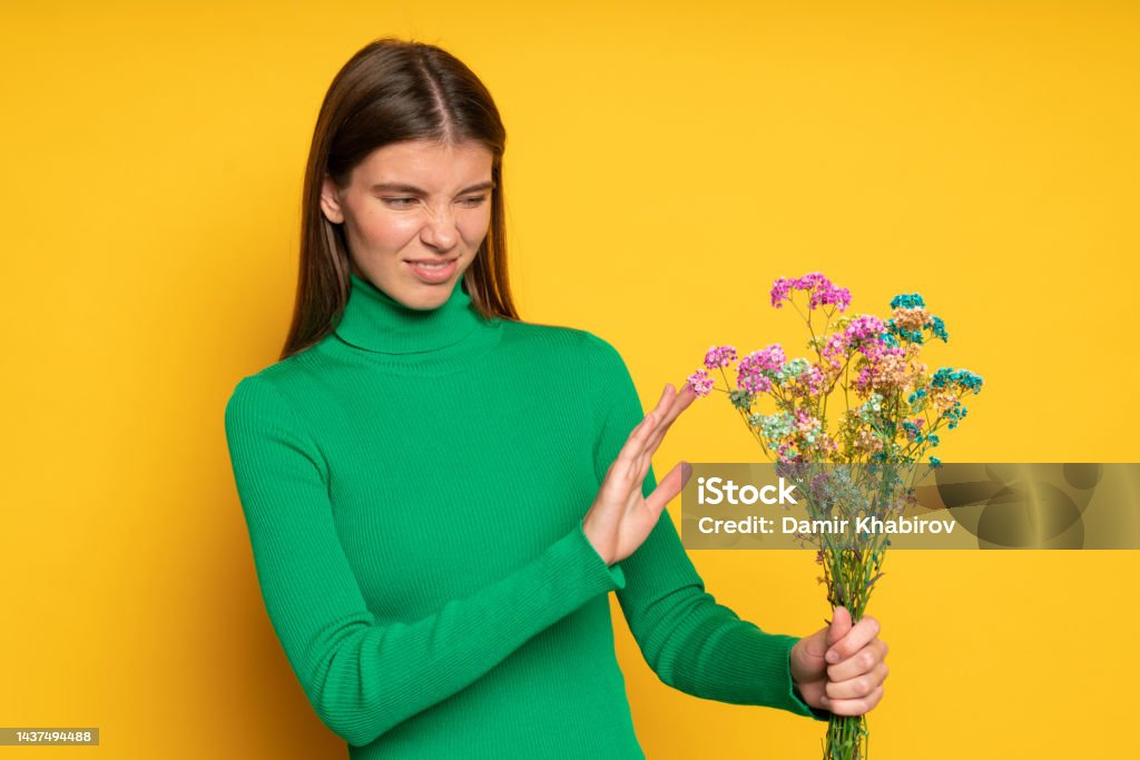 Allergic woman holding bouquet of flowers doing no gesture with ugh facial expression Portrait of displeased woman holding bouquet of flowers doing no gesture, posing on yellow studio background with ugh facial expression because of allergy to pollen or disgusting flower smell Allergy Stock Photo