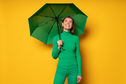 Portrait of happy joyful millennial girl in green turtleneck and jeans standing under umbrella on yellow studio background, enjoying rainy day and fall weather, looking up at cloudy sky