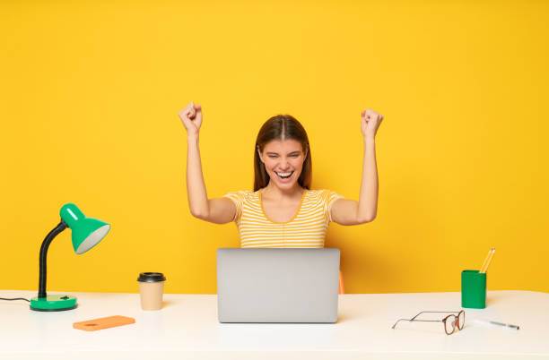 istock Student girl at table with laptop celebrating triumph with raised hands after passing exam online 1437494506