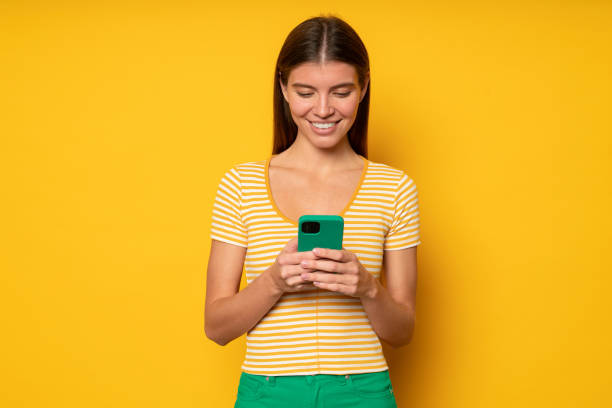 istock Woman holding phone looking at screen with smile browsing dating application on yellow background 1437494515