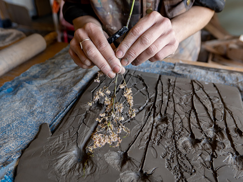Close up of the hands of a young female artist carefully lifting pressed flowers from rolled out clay. The plants have been pressed into the clay to create and imprint of favourite flowers, before pouring liquid plaster over the cast.