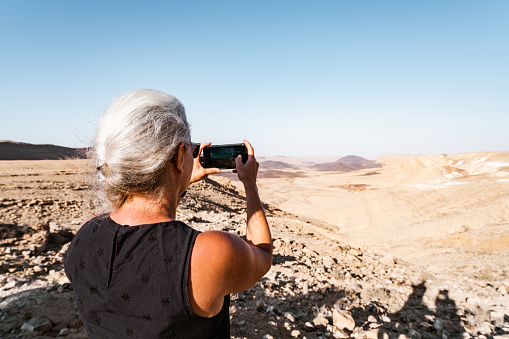 Mature woman taking photos while visiting desert in Israel. She is wearing casual clothing and sun glasses.