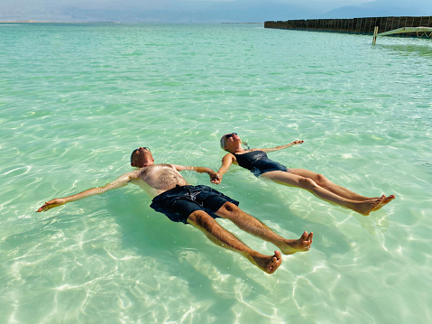 Mature couple holding hands while floating in the shallow water on the beach of Dead Sea, Israel.