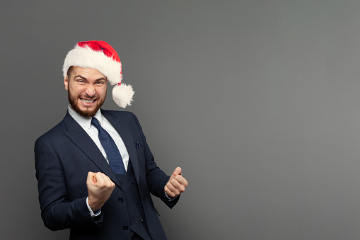 Portrait of happy excited businessman in Christmas hat laughing against grey studio wall background. Business win, fun and success concept