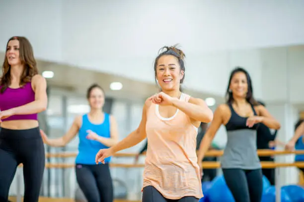A small group of mature woman stand spread apart in a fitness studio as they participate in an Aerobics class together.  They are each dressed comfortably in athletic wear and are smiling as they enjoy themselves.