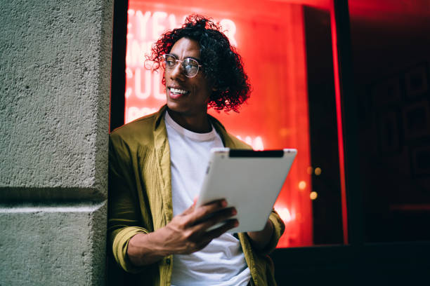 istock Smiling curly hipster guy in eyewear standing on neon light street with digital tablet share contetnt in vlog,handsome man influencer using touchpad for blogging and share media and multimedia content 1437504111