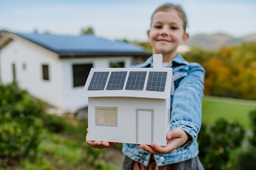 Close up of happy girl holding paper model of house with the solar panels.Alternative energy, saving resources and sustainable lifestyle concept.