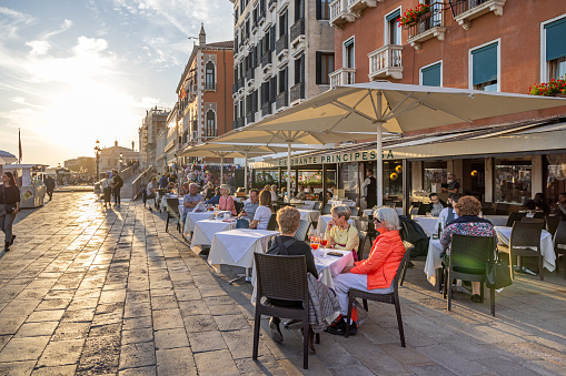 Venice, Italy - October 11th 2022: Tourists enjoy a meal at a popular outdoor restaurant in low sunlight on a public square in the center of the old and famous Italian city Venice