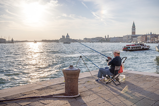 Venice, Italy - October 11th 2022: An angler sitting in a camping chair on the quay looking over the old harbor in the center of the old and famous Italian city Venice