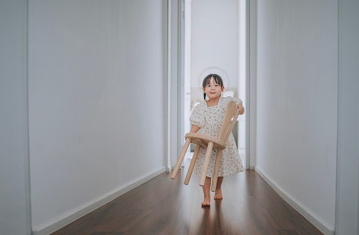 Asian Chinese young girl carrying wooden chair walking towards camera at home