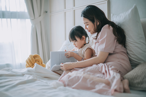 Asian Chinese pregnant mother reading book with her daughter on bed during weekend