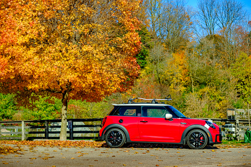 Toronto, Ontario, Canada- October 29, 2022. Chili red colour MINI COOPER on the road in public park against fall nature colours in Toronto East side, Canada. This is the third generation model F56 JCW, since BMW took over iconic brand of MINI. MINI featured in the photo is John Cooper Works model, the most powerful 2 door version. For the first time, this compact car features engine build and designed by BMW, and packs even more power and torque than previous models since 2002 to present. Original design clues and themes are still present on this brand new model. Mini has been around since 1959 and has been owned and issued by various car manufacturers.
