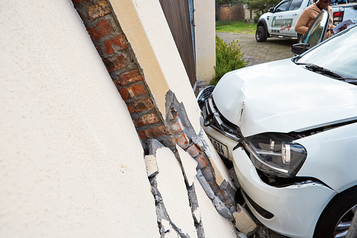 Cape Town, South Africa - August 11, 2022: The aftermath of a traffic accident between a white car and the surrounding wall of a suburban house, which has collapsed, in the southern suburbs of Cape Town, South Africa. The car is bent and buckled. A tow truck and its driver can be seen in the background.