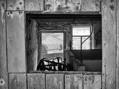 view though a broken window into an old agricultural building with peeling distressed walls and an open door with sunlit fields in the distance