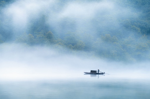 Scenic view of silhouette of man in small fishing boat reflected on surface of water on thick foggy lake during sunset on background of forest