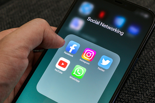 Hand holding a smartphone, clicking on screen displaying social media apps such as Facebook, Instagram, Twitter, YouTube and WhatsApp with their icons. Social networking concept.